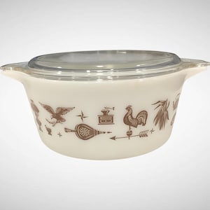 May include: Off-white Pyrex casserole dish with a clear glass lid. The dish features brown illustrations of a rooster, eagle, and other farm-themed designs. The dish has two handles and is designed for cooking and serving.