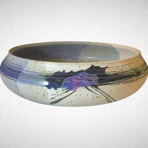 May include: A ceramic bowl with a wide, shallow design. The bowl features a speckled off-white base with blue and black painted accents. A purple floral design is painted on the side.