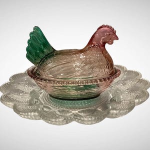 May include: A decorative glass hen-shaped dish with a matching scalloped plate. The hen is clear with a pink head and a green tail. The dish is designed to hold eggs or other small items. The plate is clear glass.