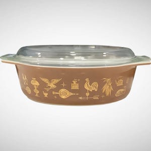 May include: A brown casserole dish with a clear glass lid. The dish features gold-colored illustrations of various objects, including a rooster, eagle, and wheat. The dish has a capacity of approximately 2 quarts.