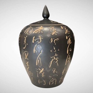 May include: A dark gray ceramic jar with a lid and a pointed finial. The jar is adorned with etched, light brown Asian characters. The jar has a rounded body and a lid that fits snugly on top. The overall design is elegant and traditional.