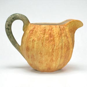 May include: A ceramic pitcher with a textured, pumpkin-like body in shades of yellow and orange. The handle is a muted green, and the spout is curved. This decorative pitcher is suitable for serving beverages.