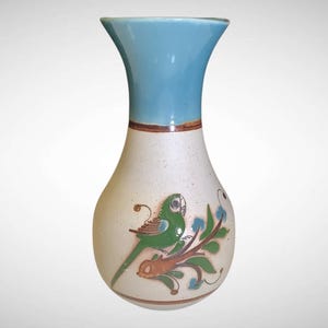 May include: A ceramic vase with a light blue top and a white body. The body features a hand-painted design of a green parrot perched on a brown branch with green leaves. The vase has a flared top and a rounded base.