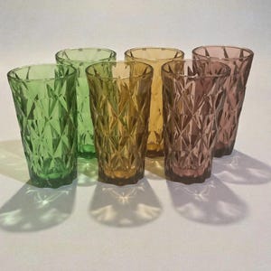 May include: Set of six colored drinking glasses. The glasses are in shades of green, amber, and pink. Each glass has a diamond pattern. The glasses are arranged in a row, with their shadows visible on the white surface.