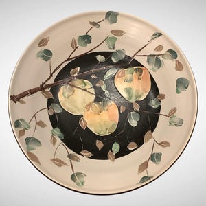 May include: A large, round ceramic serving bowl with a hand-painted design. The bowl features a cream-colored rim and a black center with three yellow and orange fruit. Brown branches and green leaves are painted around the fruit.