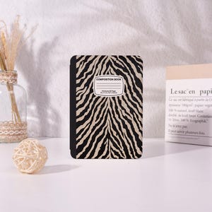 May include: A composition book with a black and beige zebra stripe pattern. The cover features a white rectangular label with the text COMPOSITION BOOK, 100 Sheets 200 Pages, and 9.75 x 7.5 in / 24.7 x 19.0 cm printed in black ink.
