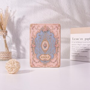 Op de afbeelding: Een rechthoekige Kindle e-reader hoes met een lichtroze en blauwe achtergrond, voorzien van gedetailleerde goudkleurige filigraan, bloemmotieven en krulwerk. Het woord KINDLE staat in een decoratief goudkleurig kader onderaan het midden.