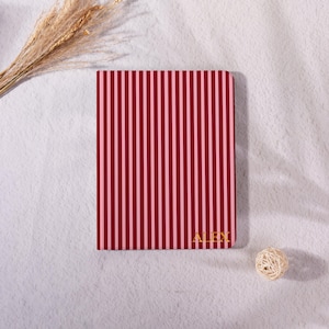May include: A notebook with a red and pink striped pattern, featuring the name "ALEX" in gold. The design has vertical stripes. The background is a white, textured surface. A small decorative ball and dried plants are also present.