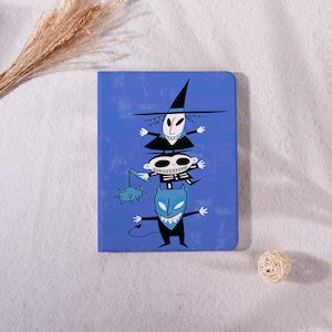 May include: A rectangular blue notebook featuring a graphic illustration of three stacked cartoon characters. The top character wears a witch hat, the middle character has a skeleton design, and the bottom character is blue with a pointed tail.