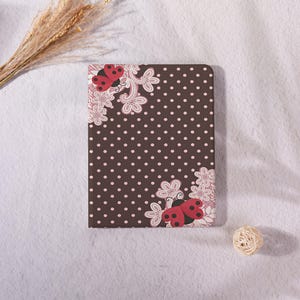 May include: A rectangular tablet case featuring a dark brown background with a repeating pattern of small white polka dots. The design includes two red ladybugs with black spots and white floral lace-like accents in the top left and bottom right corners.