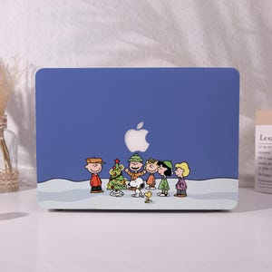 May include: A blue laptop case featuring a festive scene with cartoon characters, a Christmas tree, and a snowy landscape. The Apple logo is visible. The design includes characters in winter attire.