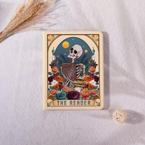 Peut inclure: Une carte de tarot avec un squelette tenant un livre, entouré de fleurs orange, violettes et bleues. La carte a une bordure crème avec le texte "THE READER". Un soleil jaune et un fond bleu sont visibles.