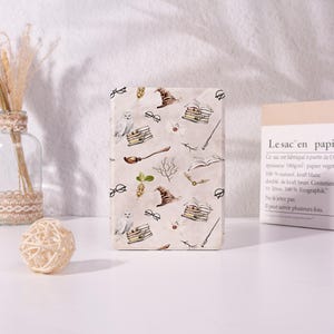 Op de afbeelding: Een rechthoekige e-readerhoes met een lichtbeige achtergrond en een herhalend patroon van illustraties in tovenaarsstijl, waaronder witte uilen, bruine sorteerhoeden, stapels boeken, ronde brillen, toverstokken, enveloppen en gouden snaaien.