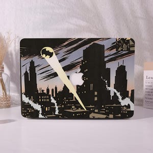 May include: A rectangular laptop skin featuring a graphic illustration of a dark city skyline at night. A bright yellow bat signal shines into the sky, and a white Apple logo is visible in the center of the design against the black buildings.