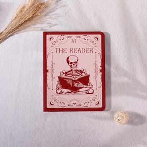 Peut inclure: Une carte de tarot rouge avec le texte "THE READER" et un squelette lisant un livre. La carte a une bordure décorative et est posée sur un fond blanc avec des plantes séchées et une boule décorative.