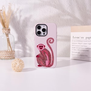 May include: A light pink smartphone case featuring a stylised illustration of a monkey in shades of dark pink and red. The monkey is depicted in a seated position with its long, curled tail extending upwards. The case has a black camera cutout.