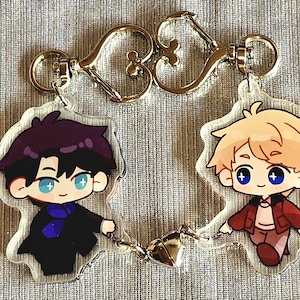 May include: Two clear acrylic keychains connected by a silver heart-shaped charm. Each keychain features a cartoon illustration of a character with black hair and a character with blonde hair. The keychains have gold-colored clasps.