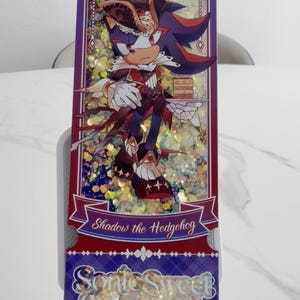 May include: A rectangular shaker charm featuring Shadow the Hedgehog in a pirate-themed outfit holding a cake. The background is filled with gold glitter, framed by a purple border with white accents. Text reads Shadow the Hedgehog and Sonic Sweet Adventure.