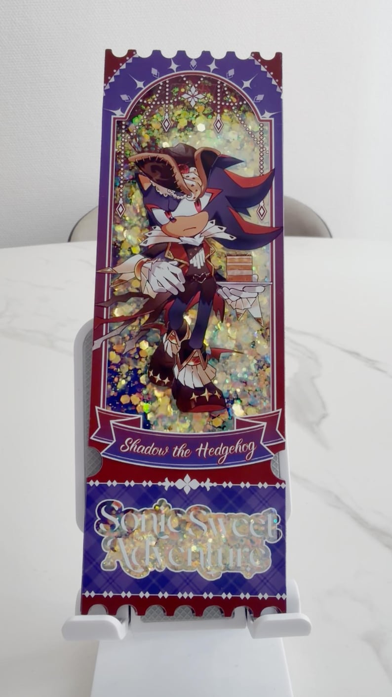 May include: A rectangular shaker charm featuring Shadow the Hedgehog in a pirate-themed outfit holding a cake. The background is filled with gold glitter, framed by a purple border with white accents. Text reads Shadow the Hedgehog and Sonic Sweet Adventure.