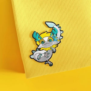 May include: An embroidered patch featuring a stylized white creature with teal accents, yellow hair, and large eyes, attached to a bright yellow ribbed fabric. The creature has a curved tail, small claws, and a floral detail near its head.