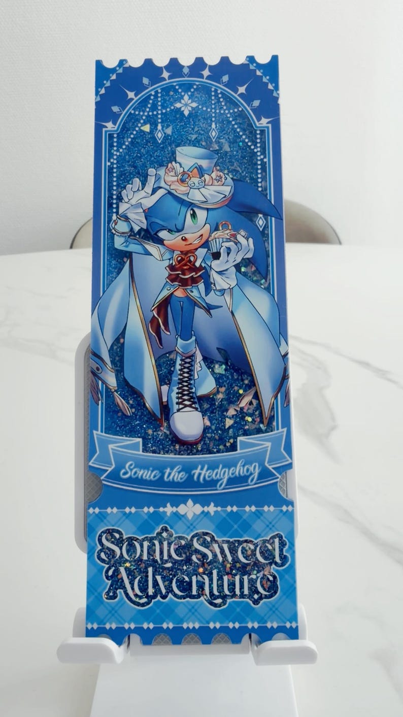May include: A rectangular blue shaker bookmark featuring an illustration of Sonic the Hedgehog wearing a formal outfit with a top hat and cape. The design includes glittery blue accents, a cupcake graphic, and the text Sonic the Hedgehog and Sonic Sweet Adventure printed in a decorative font.