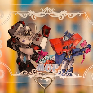 Puede incluir: Tarjeta acrílica transparente con los personajes de dibujos animados Megatron y Optimus Prime, sosteniendo lazos rosas. La tarjeta tiene un borde decorativo y la palabra "MOP". Un soporte de alambre en forma de corazón sostiene la tarjeta.
