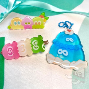 Op de afbeelding: Drie kleurrijke pinnen met cartoonfiguren. De eerste pin is een groen, geel en roze personage met een groen blaadje bovenop. De tweede pin is een groen, wit en roze personage met een groen blaadje bovenop. De derde pin is een blauw personage met een wit en blauw ontwerp.