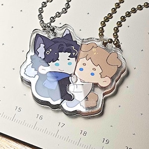 May include: Two clear acrylic keychains with cartoon illustrations of anime-style characters. One character has dark blue hair and a wolf-like appearance, the other has light brown hair. Both have blue eyes and are connected by a chain.