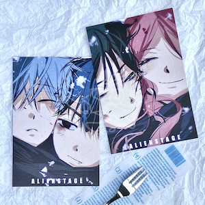 Könnte beinhalten: Zwei Kunstdrucke im Anime-Stil mit männlichen Charakteren mit blauen und rosa Haaren. Die Drucke tragen unten den Text "ALIENSTAGE". Ein kleines, durchsichtiges Plastiktütchen mit Text und eine silberne Gabel befinden sich ebenfalls im Bild.