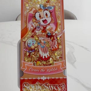 May include: A rectangular shaker charm featuring Cream the Rabbit and a Chao character. The design includes gold glitter, orange accents, and the text Cream the Rabbit and Sonic Sweet Adventure. The charm has a ticket-like shape with scalloped edges.