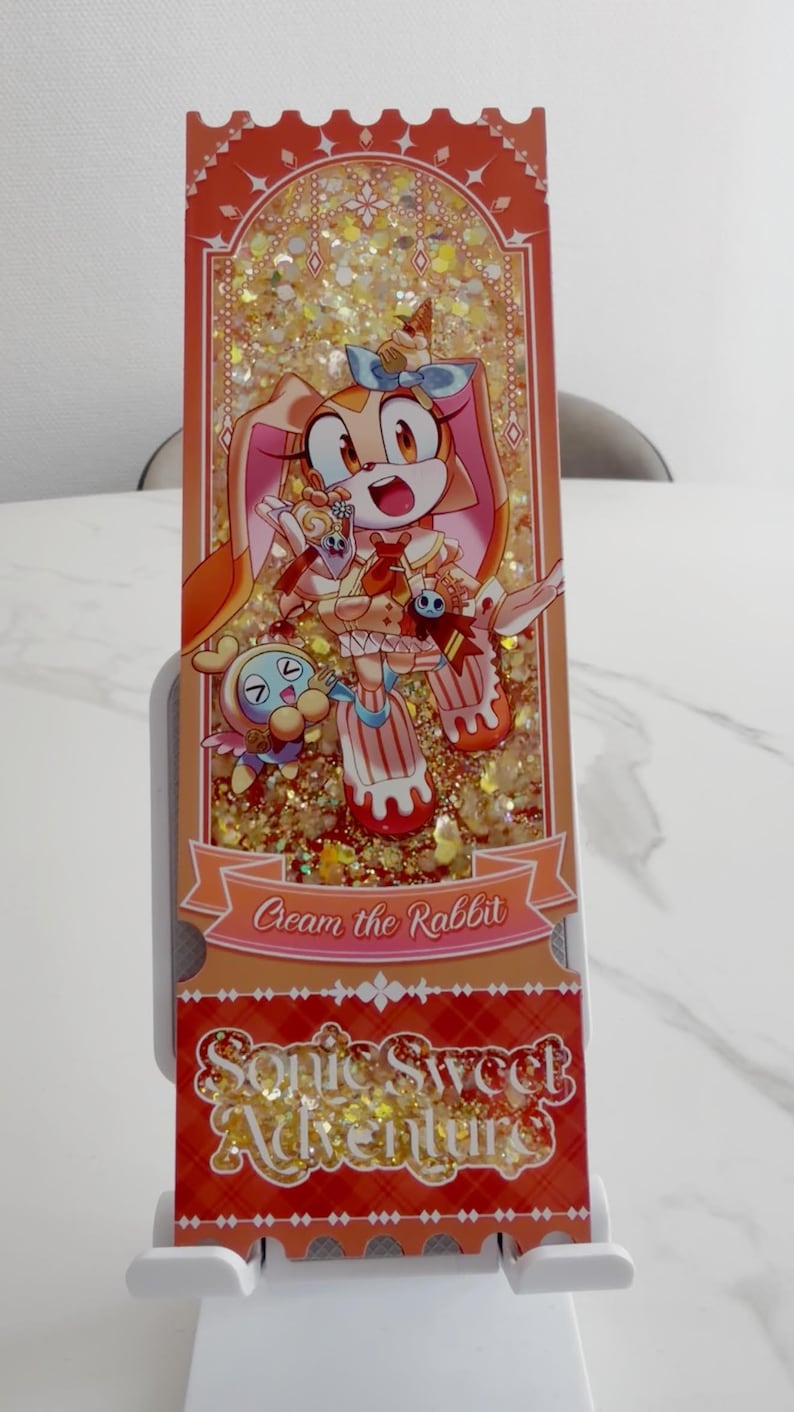 May include: A rectangular shaker charm featuring Cream the Rabbit and a Chao character. The design includes gold glitter, orange accents, and the text Cream the Rabbit and Sonic Sweet Adventure. The charm has a ticket-like shape with scalloped edges.
