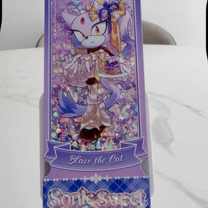 May include: A rectangular purple shaker bookmark featuring an illustration of Blaze the Cat holding a small cake. The design includes holographic glitter, a banner with the text Blaze the Cat, and a bottom section with the text Sonic Sweet Adventure.