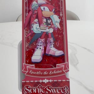 May include: A rectangular shaker bookmark featuring Knuckles the Echidna holding a small cake. The design includes red glitter, a decorative frame, and the text Knuckles the Echidna and Sonic Sweet Adventure printed in a stylized white font on a red background.