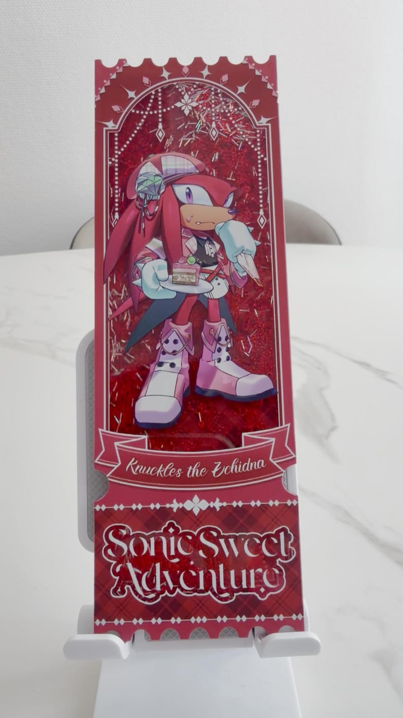 May include: A rectangular shaker bookmark featuring Knuckles the Echidna holding a small cake. The design includes red glitter, a decorative frame, and the text Knuckles the Echidna and Sonic Sweet Adventure printed in a stylized white font on a red background.