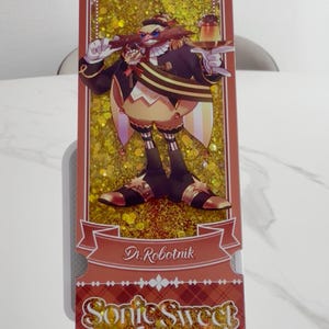 May include: A rectangular bookmark featuring a glittery gold background and an illustration of Dr. Robotnik holding a dessert. The text Dr. Robotnik appears on a banner, and the text Sonic Sweet Adventure is printed in a decorative font at the bottom.