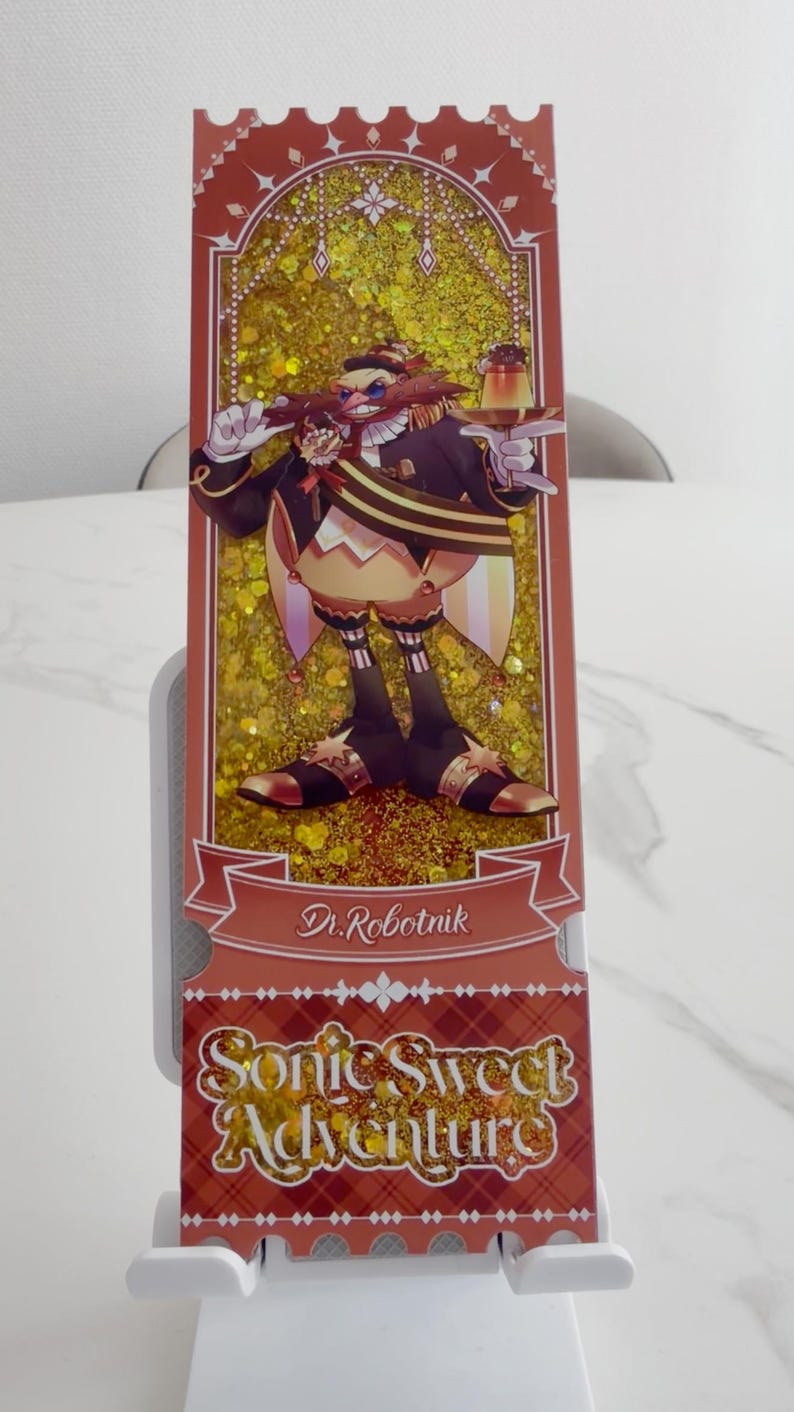 May include: A rectangular bookmark featuring a glittery gold background and an illustration of Dr. Robotnik holding a dessert. The text Dr. Robotnik appears on a banner, and the text Sonic Sweet Adventure is printed in a decorative font at the bottom.