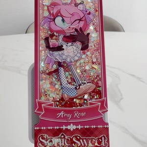 May include: A rectangular shaker bookmark featuring a pink-furred character with a wink, wearing a white apron and holding a dessert. The background is filled with iridescent glitter. The text reads Amy Rose and Sonic Sweet Adventure.