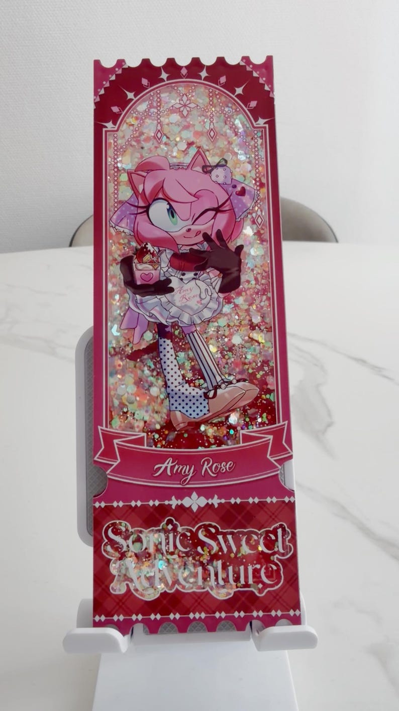 May include: A rectangular shaker bookmark featuring a pink-furred character with a wink, wearing a white apron and holding a dessert. The background is filled with iridescent glitter. The text reads Amy Rose and Sonic Sweet Adventure.