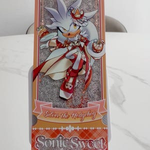 May include: A rectangular bookmark featuring an illustration of Silver the Hedgehog in a red and white outfit, holding small desserts. The design includes a peach-colored border, a plaid pattern at the bottom, and the text Silver the Hedgehog and Sonic Sweet Adventure.