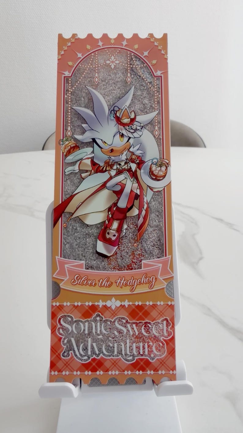 May include: A rectangular bookmark featuring an illustration of Silver the Hedgehog in a red and white outfit, holding small desserts. The design includes a peach-colored border, a plaid pattern at the bottom, and the text Silver the Hedgehog and Sonic Sweet Adventure.