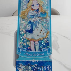 May include: A rectangular shaker bookmark featuring an anime-style illustration of Maria Robotnik in a blue and white dress holding a blue bowl. The background contains iridescent glitter. Text reads Maria Robotnik and Sonic Sweet Adventure.