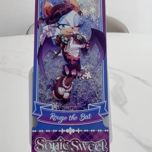 May include: A rectangular bookmark featuring an illustration of the character Rouge the Bat in a purple and white outfit. The design includes a glittery background, a blue banner with the text Rouge the Bat, and the text Sonic Sweet Adventure.