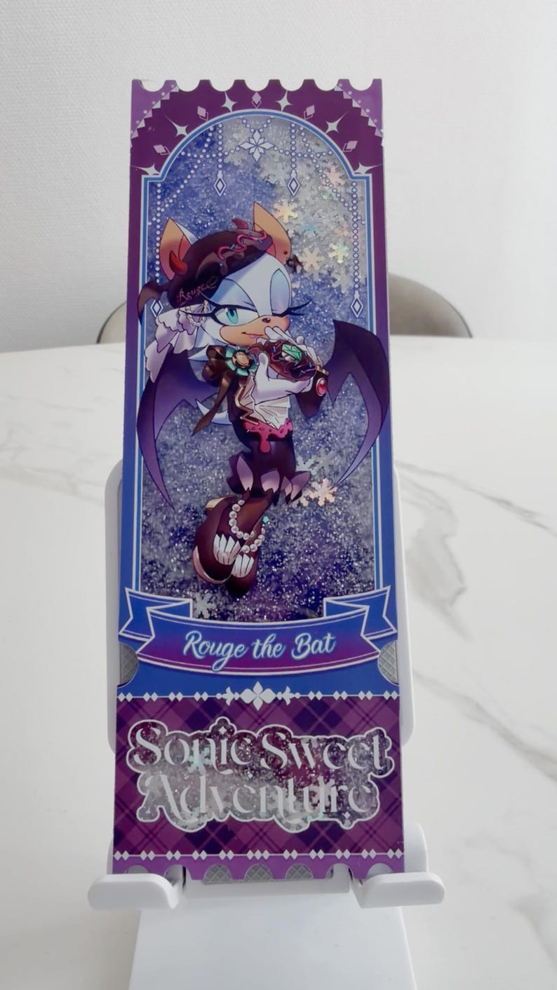 May include: A rectangular bookmark featuring an illustration of the character Rouge the Bat in a purple and white outfit. The design includes a glittery background, a blue banner with the text Rouge the Bat, and the text Sonic Sweet Adventure.