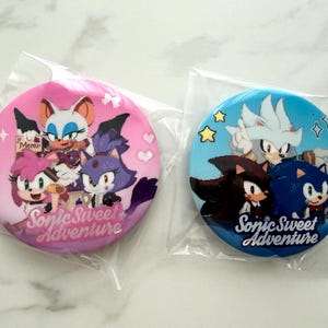 May include: Two round pinback buttons featuring characters from the Sonic the Hedgehog franchise. One button is pink with characters and the text "Sonic Sweet Adventure." The other is blue with different characters and the same text.