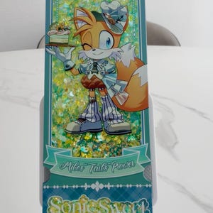 May include: A rectangular shaker bookmark featuring a cartoon fox character wearing a chef hat and outfit, holding a small cake. The background is filled with yellow and green glitter. Text reads Miles Tails Prower and Sonic Sweet Adventure.