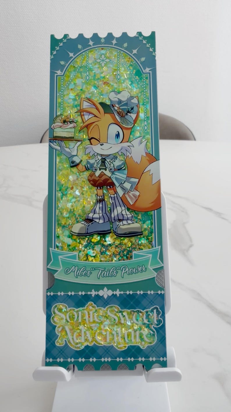 May include: A rectangular shaker bookmark featuring a cartoon fox character wearing a chef hat and outfit, holding a small cake. The background is filled with yellow and green glitter. Text reads Miles Tails Prower and Sonic Sweet Adventure.