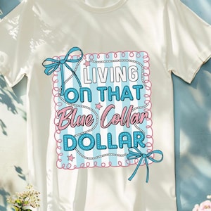 May include: White t-shirt with a graphic that reads "LIVING ON THAT Blue Collar DOLLAR" in a blue and pink design. The design includes bows, stars, and a striped background. The shirt has a crew neck and short sleeves.