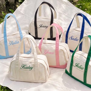 May include: Six off-white canvas duffel bags with colored trim and handles. Each bag features an embroidered name in a matching color: Bella in light blue, Jenny in black, Sadie in royal blue, Kaley in dark green, Grace in pink, and Julia in green.