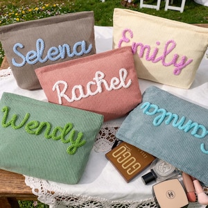 May include: Five corduroy cosmetic bags in various colors: taupe, peach, pale yellow, green, and teal. Each bag has a name embroidered in a contrasting color: Selena, Rachel, Emily, Wendy, and Camila. The bags are displayed on a white lace tablecloth.