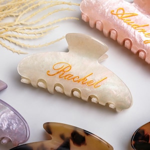 May include: Several personalised hair claw clips in different colours. The main clip is off-white with the name "Rachel" in gold script. Other clips include pink and tortoiseshell patterns, each with a name.